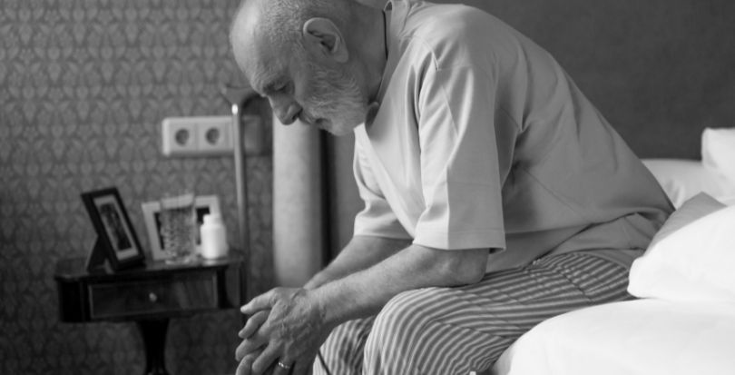 Sad elderly man sitting leaned over on side of the bed